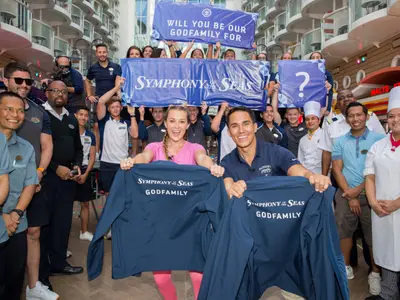 Ein Mann und eine Frau halten dunkelblaue Jacken mit dem Logo 'Symphony of the Seas Godfamily'. Sie lächeln und sind von einer Gruppe von Crewmitgliedern und anderen Personen umgeben. Im Hintergrund sind Teile der Symphony of the Seas und Banner zu sehen, die auf sie hinweisen. Die Szene wirkt festlich und fröhlich.