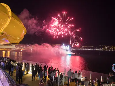 Nächtliche Szene vom Deck der Symphony of the Seas: Passagiere beobachten ein großes Feuerwerk über dem Wasser, das sich rot im Meer spiegelt. Eine beleuchtete Küstenlinie ist im Hintergrund zu sehen.