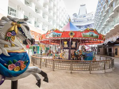 Ein farbenfrohes Karussell mit geschnitzten Tieren wie Pferden und Tigern auf der großen Symphony of the Seas. Im Hintergrund sind die weißen Balkonkabinen und der Schiffsname 'Symphony of the Seas' zu sehen. Wenige Personen sind auf der Promenade unterwegs.