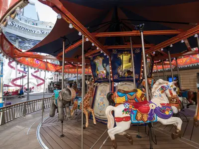 Ein buntes Karussell mit Pferden, Elefanten und Giraffen steht auf dem Promenade-Deck der Symphony of the Seas. Im Hintergrund sind Schiffsbalkone und eine rote Wasserrutsche zu sehen.