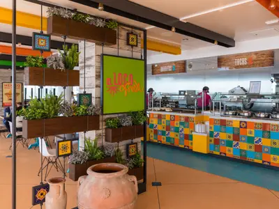 Interieur des mexikanischen Restaurants 'El Loco Fresh' auf der Symphony of the Seas. Eine bunte Theke mit warmen Speisen, Personal und hängenden Pflanzen ist sichtbar.