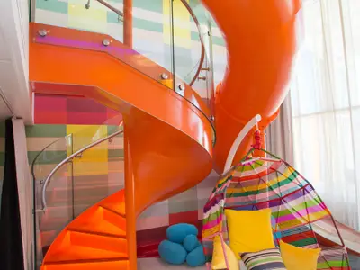 Bunte Familienkabine mit leuchtend oranger Wendeltreppe und passender Röhrenrutsche. Ein mehrfarbiger Hängesessel mit gelbem Kissen steht im Vordergrund. Die Wand ist kariert.