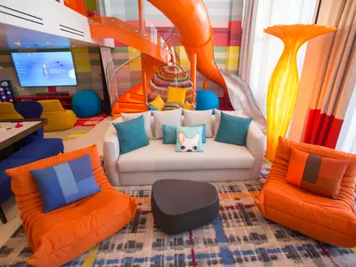 Ein buntes, zweistöckiges Spielzimmer an Bord der Symphony of the Seas mit einer orangefarbenen Rutsche, Airhockeytisch, großer Leinwand und vielfältigen Sitzgelegenheiten.