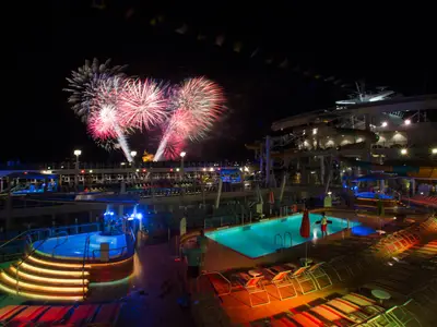 Nächtliche Szene auf dem Oberdeck der Symphony of the Seas. Beleuchtete Pools, Wasserrutschen und Liegebereiche sind zu sehen. Ein farbenprächtiges Feuerwerk erhellt den Himmel über dem Schiff.
