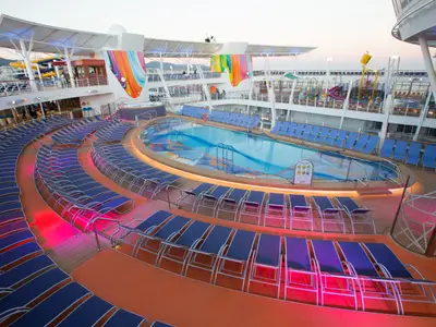 Das abendliche Pooldeck der Symphony of the Seas zeigt Reihen blauer Liegestühle um einen beleuchteten Pool. Im Hintergrund sind Wasserrutschen und farbige Deckdekorationen sichtbar.