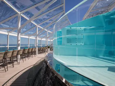 Sonniger Loungebereich an Bord der Symphony of the Seas mit einem transparenten Pool, Sitzgelegenheiten unter einem Glasdach und Meerblick.