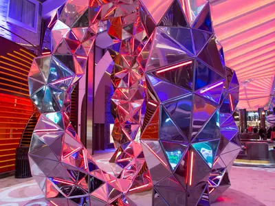 Moderne, spiegelnde Skulptur aus facettenreichen Elementen mit neonroten Lichtern im Inneren der Symphony of the Seas. Sie reflektiert die bunte Beleuchtung der Schiffspromenade.