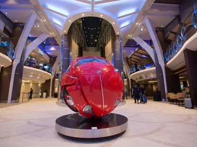 Atrium der Symphony of the Seas. Rote, kugelförmige VW Käfer Skulptur steht zentral. Mehrere Ebenen mit Geschäften und Cafés, belebt von Passagieren unter heller Beleuchtung.