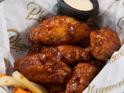 Nahaufnahme von mehreren glänzend glasierten Chicken Wings, die in einer weißen Papierschale liegen. Die Wings sind appetitlich braunrot und haben eine leicht glänzende, klebrige Sauce. Im Hintergrund ist leicht ein goldenes Logo auf dem Papier erkennbar. Ein schwarzer Rand einer Schale ist oben rechts zu sehen.