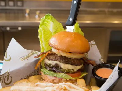 Nahaufnahme eines hohen Burgers in einer Papiermanschette. Er hat zwei Patties, geschmolzenen Käse, knusprigen Bacon, Zwiebelringe, Salat, Tomate, Gurken und rote Zwiebeln. Ein Steakmesser steckt im oberen Brötchen. Dazu eine kleine Schale Dip.