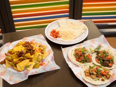Teller mit mexikanischen Gerichten auf dunklem Holztisch. Nachos mit Käse und Fleisch. Tacos mit Salsa. Quesadillas mit Tomaten. Bunte, gestreifte Wand im Hintergrund.