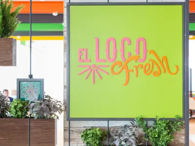 Der Schriftzug 'EL LOCO Fresh' in leuchtendem Pink und Orange auf einer lindgrünen Wand. Dekorative Pflanzen in Holzkästen und ein gemaltes Mandala sind ebenfalls sichtbar.