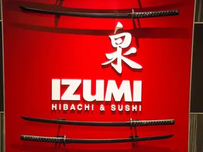 Das rote Leuchtschild des japanischen Restaurants Izumi Hibachi & Sushi mit weißem Schriftzug und einem japanischen Schriftzeichen ist zu sehen. Drei Samuraischwerter sind als Dekoration angebracht.