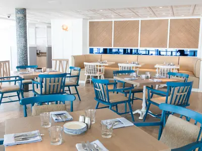 Heller Restaurantsaal der Symphony of the Seas mit Holztischen, blauen Stühlen und beiger Sitzbank. Maritime Einrichtung mit Mosaiksäule. Tische sind gedeckt.
