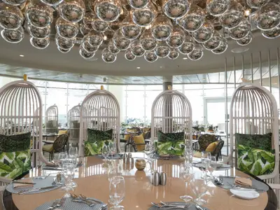 Elegantes Restaurant auf der Symphony of the Seas. Gedeckte Tische, einzigartige vogelkäfigähnliche Stühle mit grünen Kissen und reflektierende Kugeln an der Decke. Große Fensterfront.