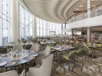Ein helles, weitläufiges Restaurant auf der Symphony of the Seas mit einer hohen, gebogenen Glasfront und einer wellenförmigen Decke. Zahlreiche Tische mit bequemen Stühlen sind festlich gedeckt. Im Hintergrund sind auffällige, vogelkäfigähnliche Sitzbereiche und eine obere Etage zu sehen. Das Ambiente ist modern und einladend.