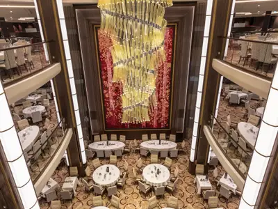 Blick von oben in den mehrstöckigen Speisesaal der Symphony of the Seas mit zahlreichen gedeckten Tischen und einem großen, goldenen Kronleuchter.