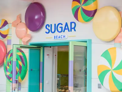 Der farbenfrohe Eingang des Süßwarenladens Sugar Beach auf der Symphony of the Seas ist mit riesigen, bunten Lollis und Bonbons dekoriert. Die Türen sind mintgrün und violett gerahmt.