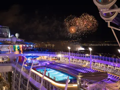 Die Symphony of the Seas bei Nacht. Gäste auf dem beleuchteten Pooldeck beobachten ein Feuerwerk über der fernen Küstenstadt.