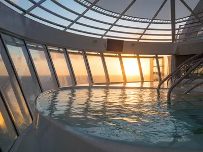 Innenpool auf der Symphony of the Seas. Goldenes Sonnenuntergangslicht durchflutet große Fenster mit Meerblick und spiegelt sich im Poolwasser. Markante Dachkonstruktion sichtbar.