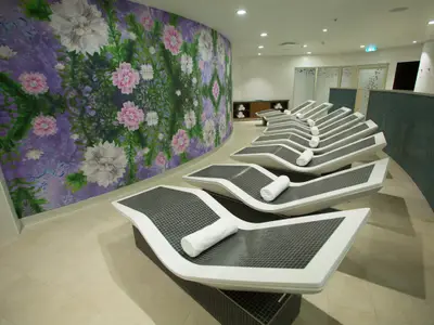 Symphony of the Seas: Entspannungsbereich mit modernen Liegen und floralem Wandbild in Pink, Weiß und Lila an einer gekrümmten Wand.