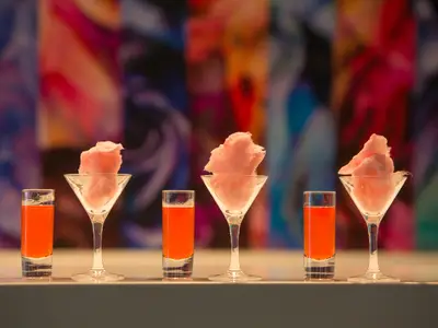 Eine Reihe von sechs Cocktailgläsern und Shotgläsern auf einer Bar. Jedes zweite Glas ist ein Martiniglas gefüllt mit rosa Zuckerwatte, dazwischen stehen Shotgläser mit orangefarbener Flüssigkeit. Der Hintergrund ist unscharf und farbenfroh.
