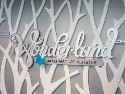 Ein Nahaufnahme-Foto des "Wonderland Imaginative Cuisine"-Schildes. Der Schriftzug "Wonderland" ist in weißer, geschwungener Schrift auf einem silbernen Hintergrund mit abstrakten, weißen Baumstrukturen zu sehen. Ein leuchtend blauer Schmetterling ziert den Schriftzug, und darunter steht auf einem grauen Balken "IMAGINATIVE CUISINE" in schwarzen Großbuchstaben.