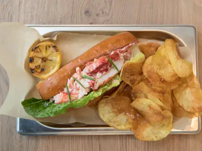 Ein appetitlich angerichtetes Hummerbrötchen mit Salat und Kräutern, begleitet von knusprigen Kartoffelchips und einer gegrillten Zitronenhälfte, serviert auf Backpapier in einem Metalltablett auf einem Holztisch.
