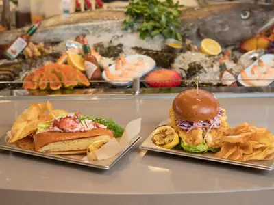 Ein Hummerbrötchen und ein Fischsandwich mit Chips und Zitrone auf zwei Tellern. Im Hintergrund eine Meeresfrüchte-Auslage auf Eis.