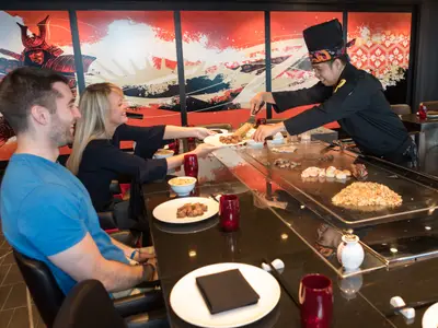 Ein Koch bereitet in einem japanischen Teppanyaki-Restaurant Essen auf einer heissen Platte zu, während Gäste zusehen und Speisen erhalten. Asiatische Wandmalerei im Hintergrund.