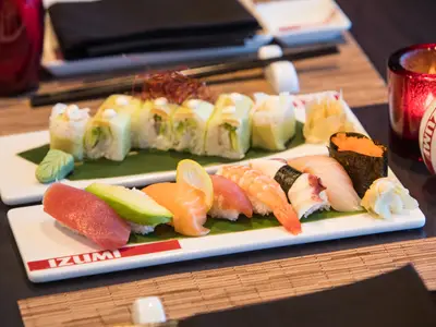 Eine weiße Platte mit frischem Sushi und Nigiri, darunter Thunfisch, Lachs, Garnelen, Oktopus, Uramaki und Gunkan Maki. Dazu Wasabi, Ingwer und Zitrone, auf einem Holztisch mit roter Dekoration.