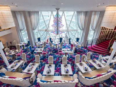 Der farbenfrohe Speisesaal auf der Symphony of the Seas zeigt große Fenster, einen floral gemusterten Teppich in Blau und Pink, sowie gedeckte Tische und verschiedene Sitzgelegenheiten.