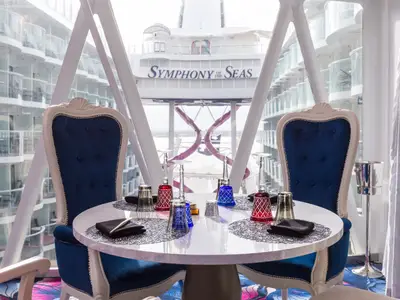 Ein elegant gedeckter Tisch für zwei Personen in einem Restaurant auf der Symphony of the Seas. Blaue Samtstühle und eine weiße Tischplatte sind zu sehen. Durch die Fenster blickt man auf die Balkone des Schiffs und den Schriftzug 'Symphony of the Seas'.