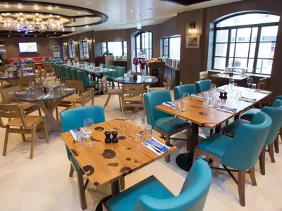 Ein modernes Restaurant auf der Symphony of the Seas mit vielen gedeckten Holztischen, tealfarbenen Stühlen und großen Fenstern. Zwei Mitarbeiter sind im Hintergrund zu sehen.