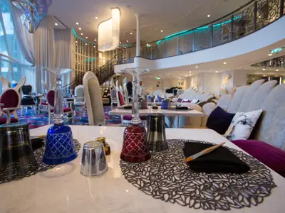 Blick in ein modernes Spezialitätenrestaurant mit blauen und roten Wassergläsern auf einem weißen Tisch im Vordergrund. Im Hintergrund sind plüschige Sessel, eine Treppe und große Fenster sichtbar.