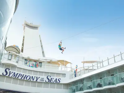 Eine Person in Sportkleidung saust an einem Seil befestigt über das Oberdeck der Symphony of the Seas. Heller, blauer Himmel über ihr.