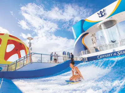 Eine junge Frau surft auf dem FlowRider Simulator an Bord der Symphony of the Seas. Im Hintergrund sind der iFLY Skydiving-Tunnel und weitere Attraktionen des Schiffes zu sehen.
