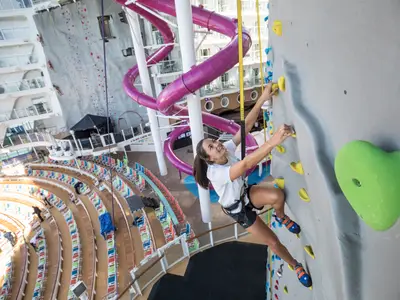 Auf der Symphony of the Seas klettert eine Frau an einer Felswand. Eine markante lila Wasserrutsche, Zuschauerbereiche und weitere Decks sind im Hintergrund zu erkennen.