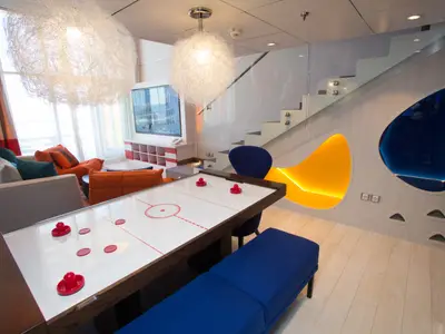 Ansicht einer modernen Suite auf der Symphony of the Seas. Mit Air-Hockey-Tisch, Wohnbereich, Treppe und farbigen Leuchtnischen.