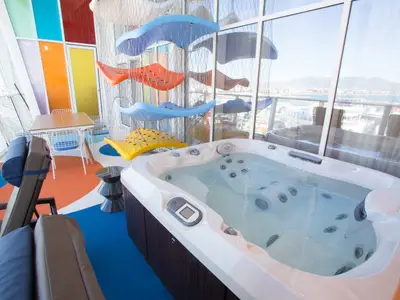 Ein Whirlpool auf Deck der Symphony of the Seas. Bunte Dekorationen, Sitzmöglichkeiten und große Fenster bieten Meerblick auf eine Hafenstadt.