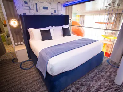 Blick in eine moderne Kabine der Symphony of the Seas mit blauem Doppelbett. Eine orangefarbene Rutsche und bunte Dekoration sind im Hintergrund durch eine Brüstung sichtbar.