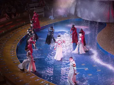 Eine Wassershow im AquaTheater der Symphony of the Seas. Künstler in roten, schwarzen und weißen Kostümen bewegen sich im Wasser, das spritzt. Zuschauer sind auf den Rängen zu sehen.