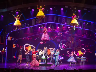 Eine lebendige Bühnenszene des Musicals Hairspray auf der Symphony of the Seas zeigt Darsteller in bunten Kostümen, teils schwebend, vor einer retro-inspirierten Kulisse mit hellen Lichtern und dem Schriftzug 'Corny Collins Show'.