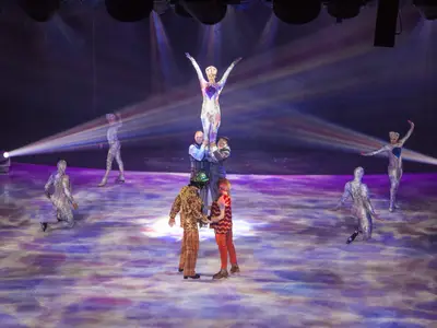 Eine farbenfrohe Eisshow auf der Symphony of the Seas. Mehrere Künstler in glänzenden Kostümen vollführen akrobatische Figuren auf dem Eis, beleuchtet von Scheinwerfern.