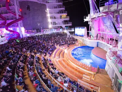 Das AquaTheater der Symphony of the Seas bei Nacht, mit Publikum, beleuchteter Poolbühne, Leinwand, Kletterwand und Wasserrutsche.