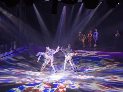 Eine farbenfrohe Eisshow auf der Symphony of the Seas. Zwei Paare in hellen Kostümen schweben über das Eis, während Scheinwerfer bunte Muster projizieren. Weitere Darsteller im Hintergrund.