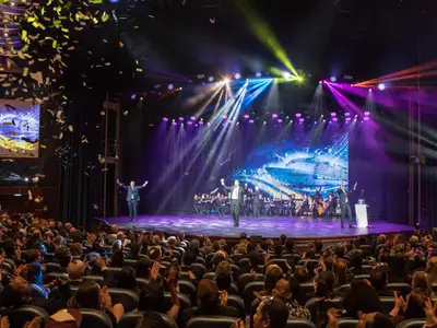 Applaudierendes Publikum im Auditorium. Auf der Bühne: Sänger, Orchester und Leinwand mit der Symphony of the Seas. Bunte Bühnenlichter, Konfetti.