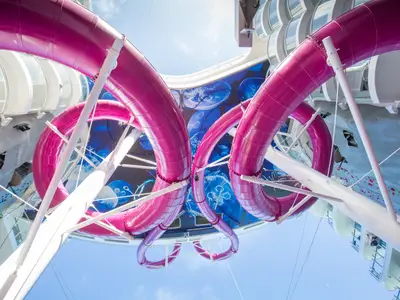 Blick nach oben auf die zwei markanten, magenta-farbenen Wasserrutschen der Symphony of the Seas. Darüber erstreckt sich eine blaue Decke mit leuchtenden Quallenmotiven. Seitlich sind Teile der Schiffsstruktur und der blaue Himmel sichtbar.