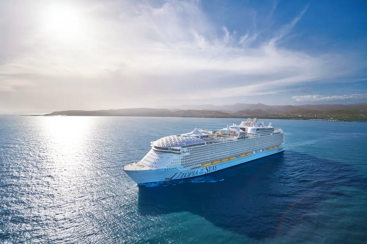 Eine Drohnenansicht zeigt die megagroße Utopia of the Seas auf blauem Meer. Im Hintergrund sind grüne Hügel und Berge unter hellem Himmel sichtbar.
