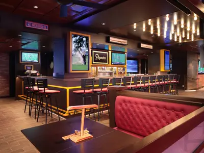 Die Playmakers Sports Bar auf der Utopia of the Seas. Eine Bar mit Hockern, rote Sitzbänke und mehrere Bildschirme zeigen Sport. Moderner Innenbereich.